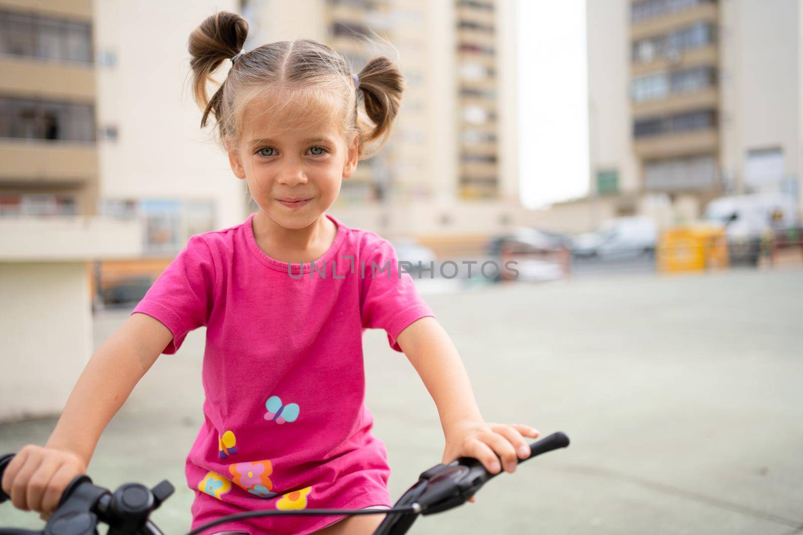 Cute little smiling girl riding bike bicycle in city on parking sunny summer day. Active family leisure with kids. Little caucasian girl 5 years old have fun with pink child bicycle