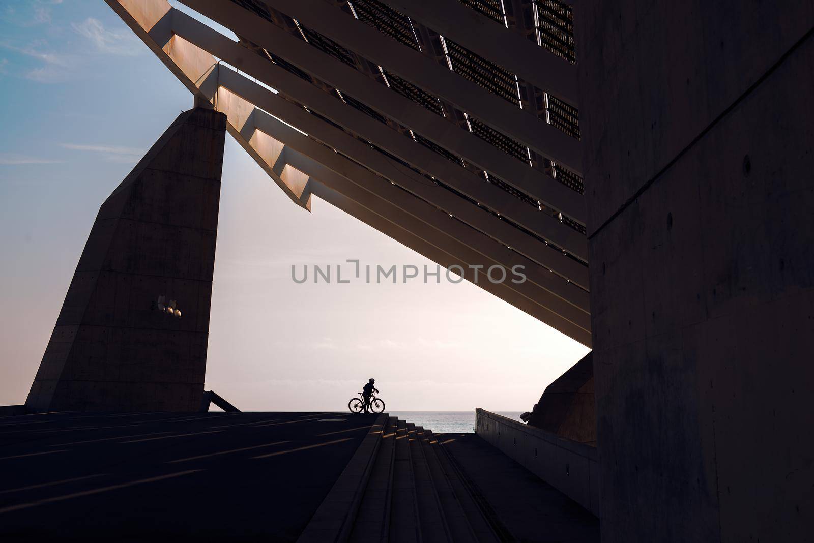 silhouette of a cyclist under a very famous steel and concrete structure of the Forum of Barcelona. The biker is next to some stairs and in the background you can see the sea, the sky is clear