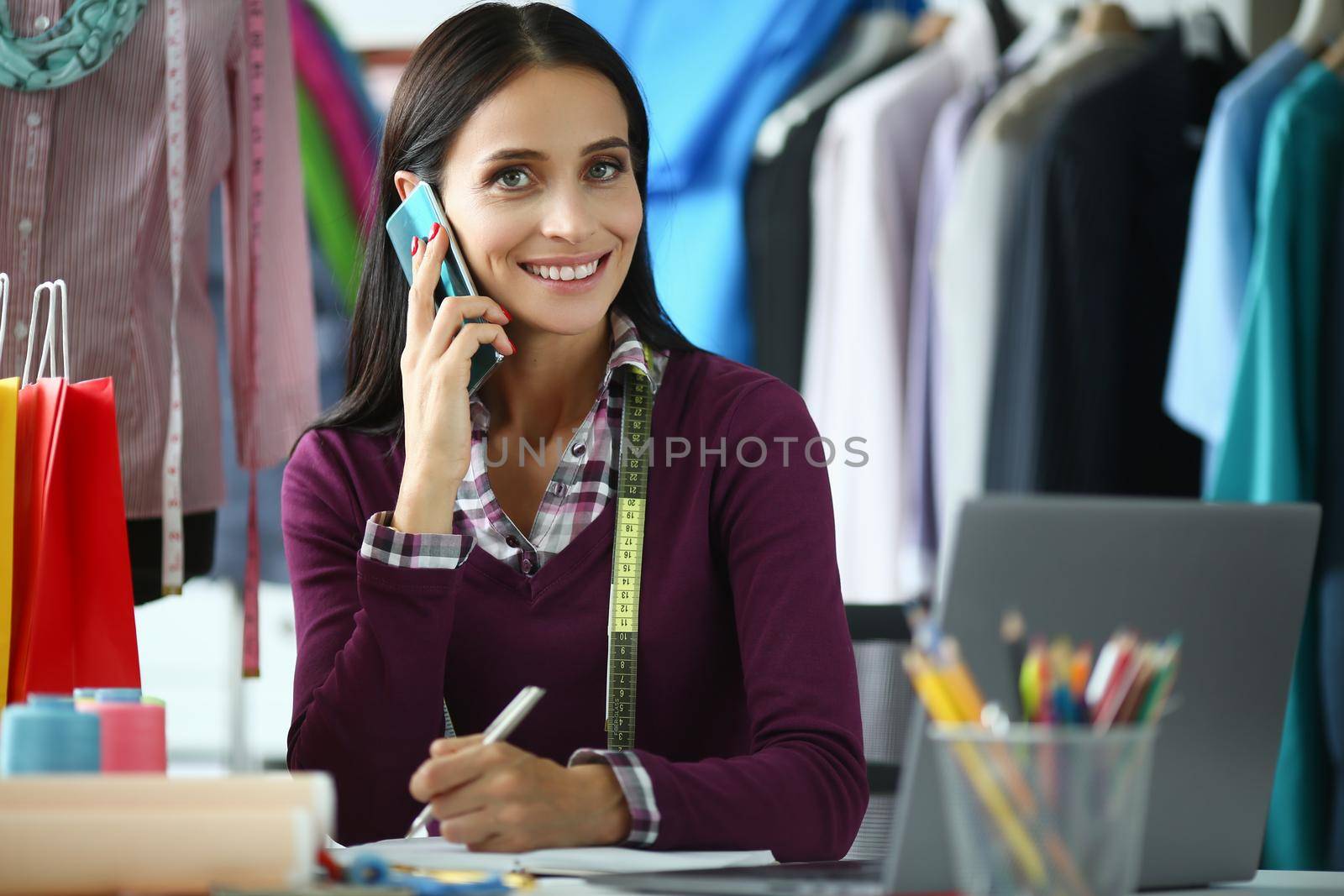 Attractive experienced seamstress needlewoman at workplace in atelier calling to client. Fashion stylist designer discussing on smartphone individual design of fabric clothing at workplace