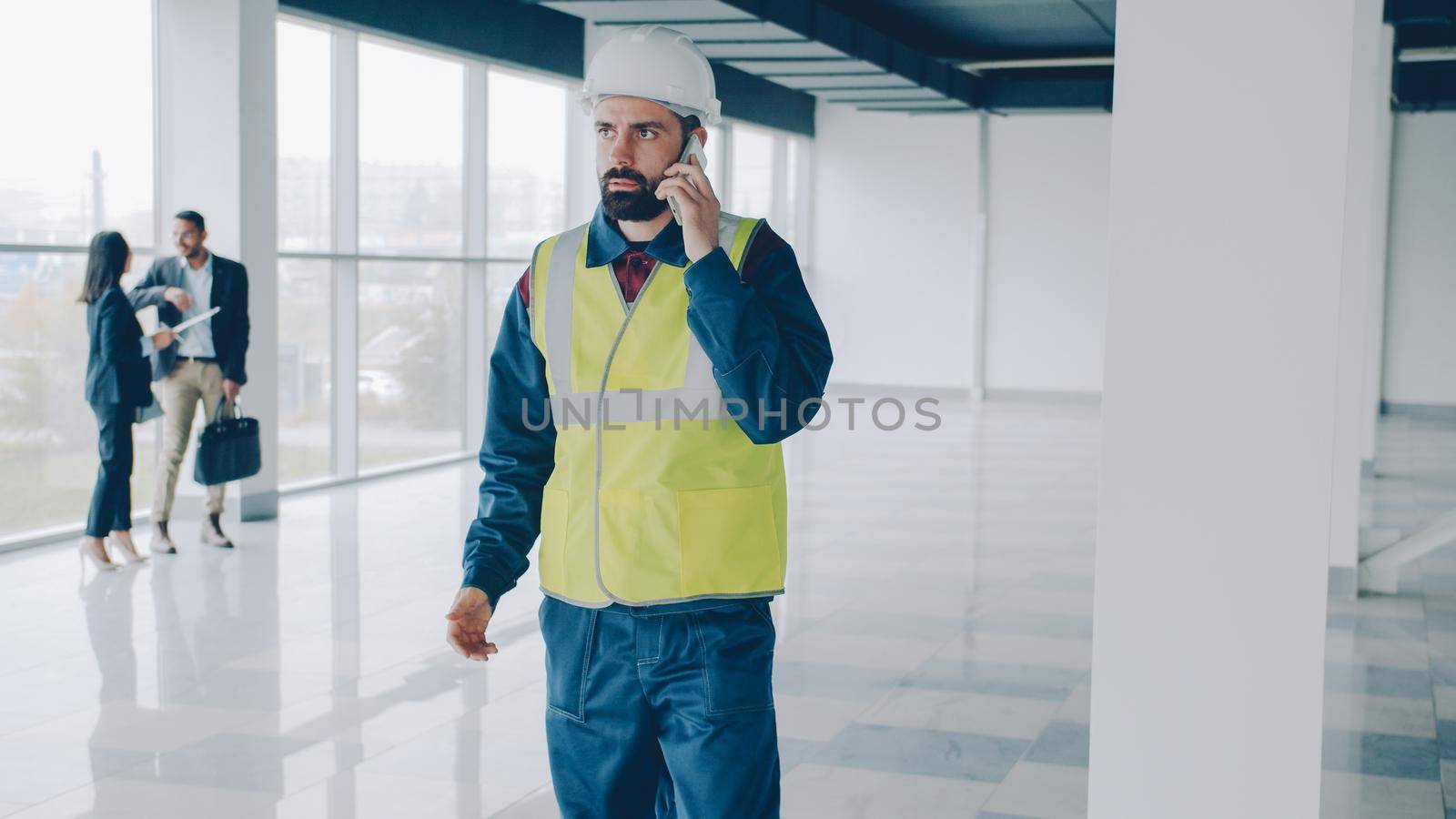 portrait of engineer wearing helmet and vest speaking on mobile phone in new building walking in empty hall concentrated on conversation