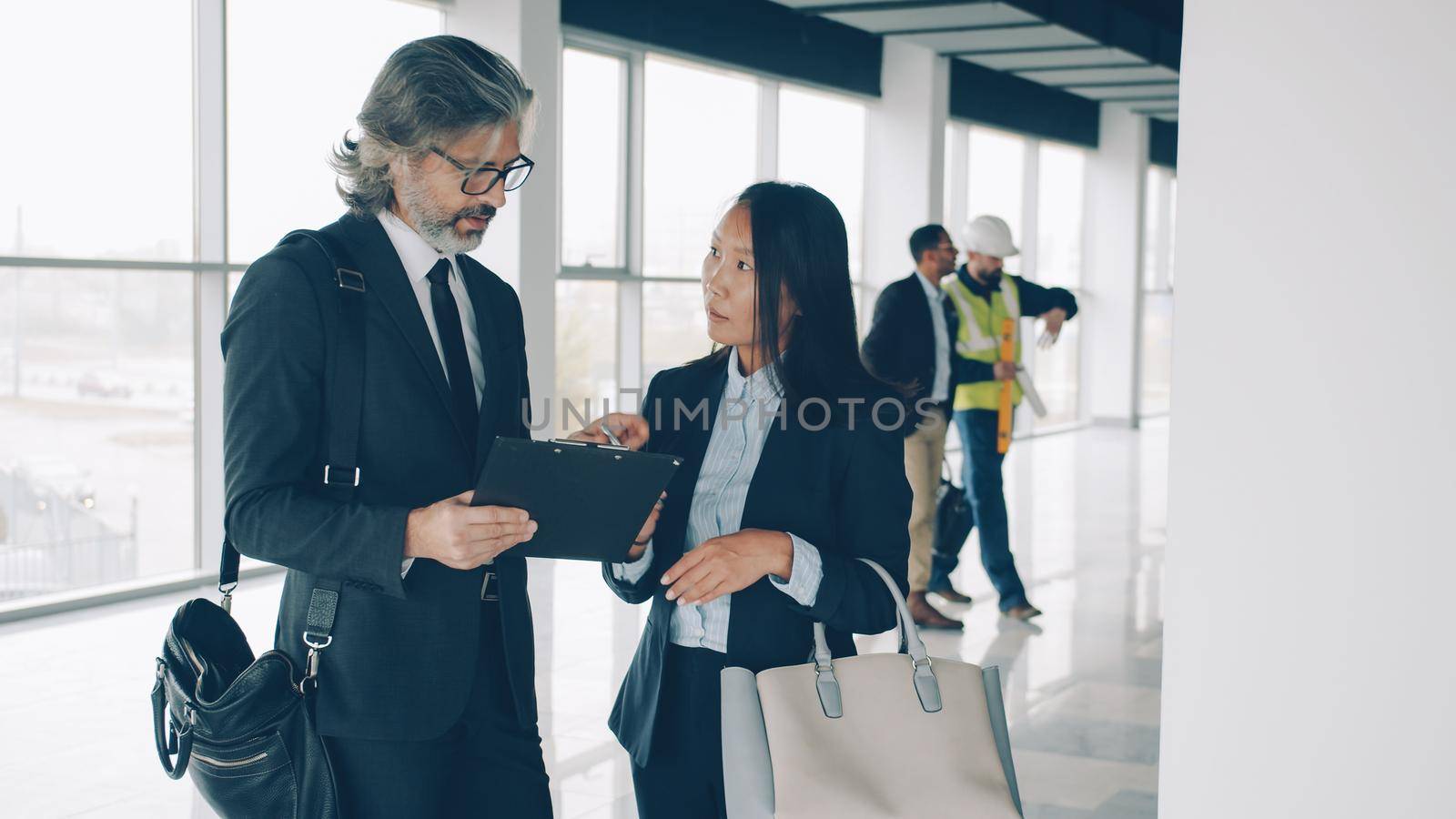 Multi-ethnic business partners man and woman are talking in modern building while workers in uniform are busy in background. People and communication concept.