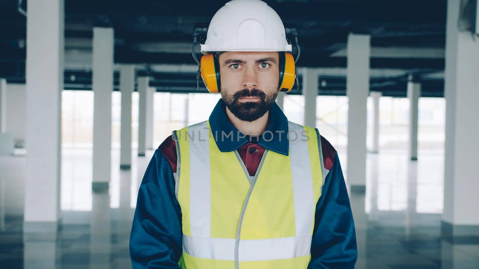 portrait of construction worker wearing safety helmet and headphones indoors in new empty industrial building. People and profession concept.