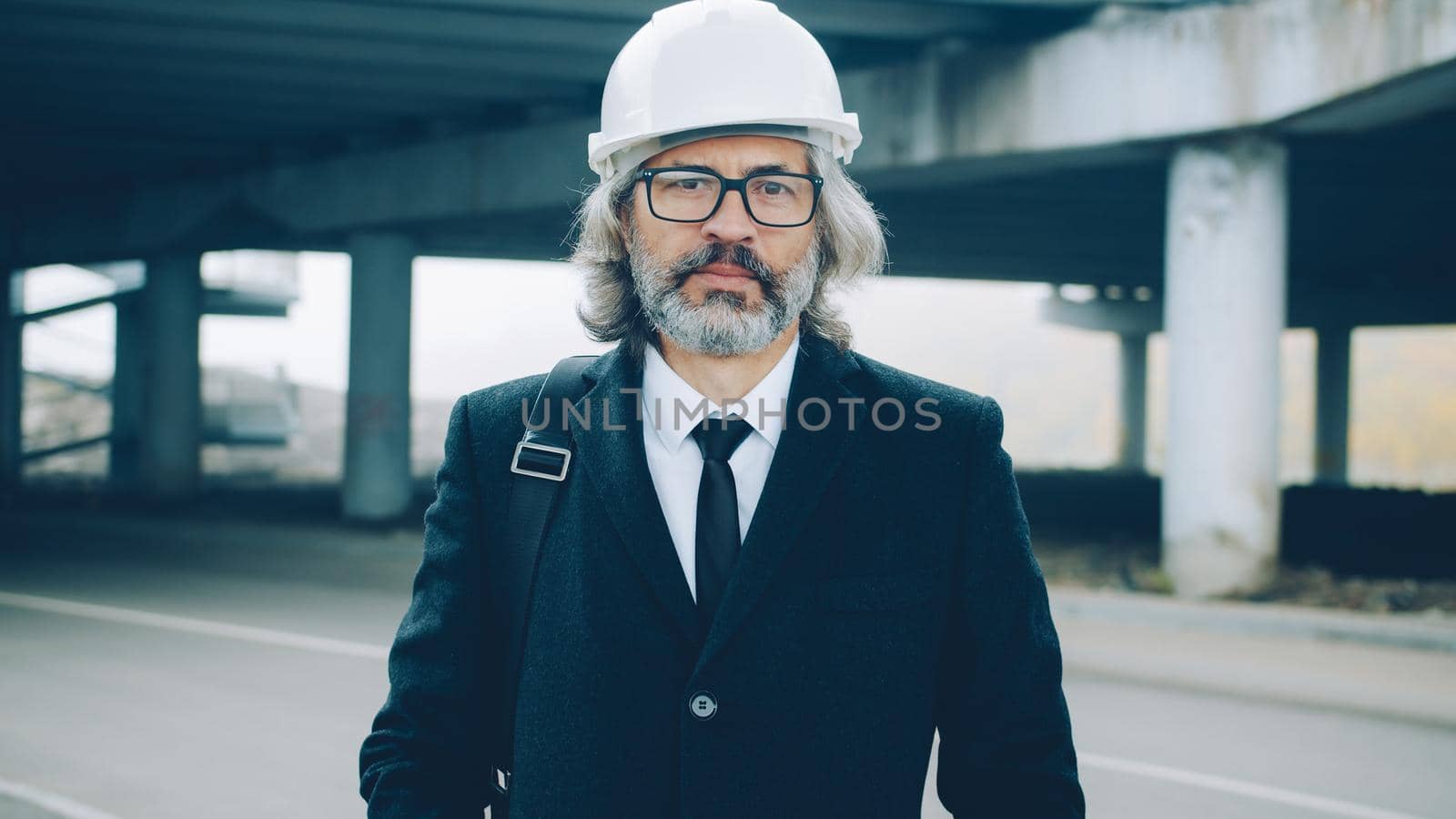 Portrait of respectful businessman standing in industrial zone outdoors wearing safety helmet and stylish suit and looking at camera with serious face