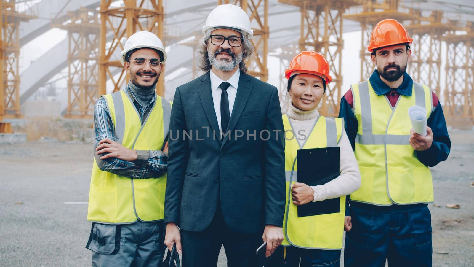 Diverse group of people building team are standing outdoors in construction site smiling looking at camera wearing helment and safety vests