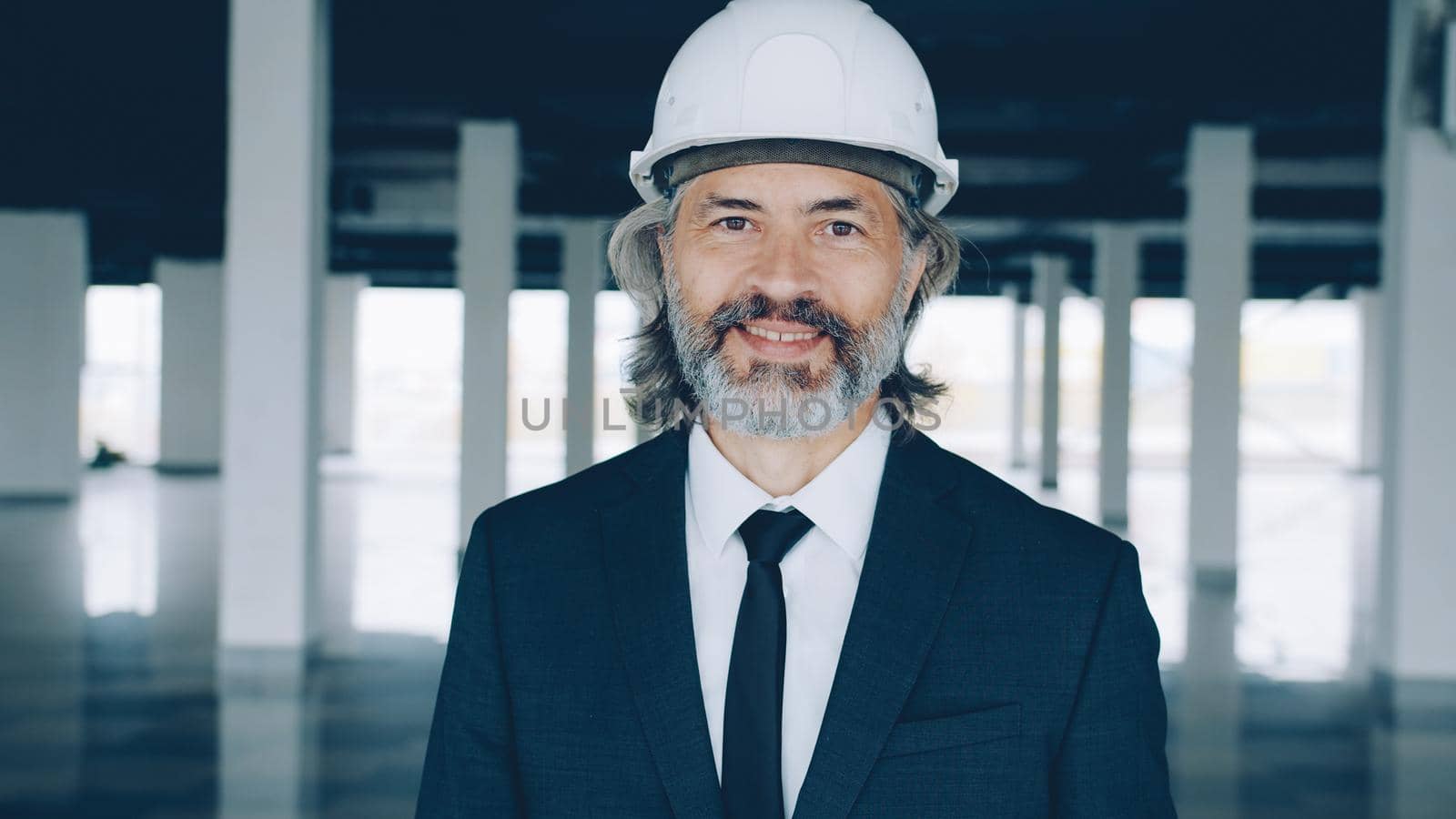 Portrait of successful real estate investor standing indoors in modern empty building wearing safety helmet and formal suit. People and occupation concept.