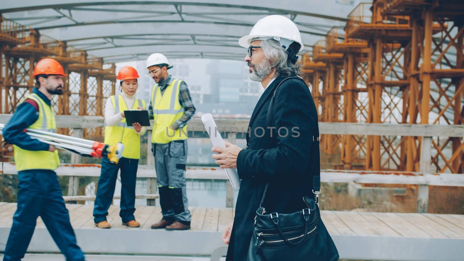 Confident mature man investor is working in construction site holding blueprint while workers are busy in background. People and business concept.