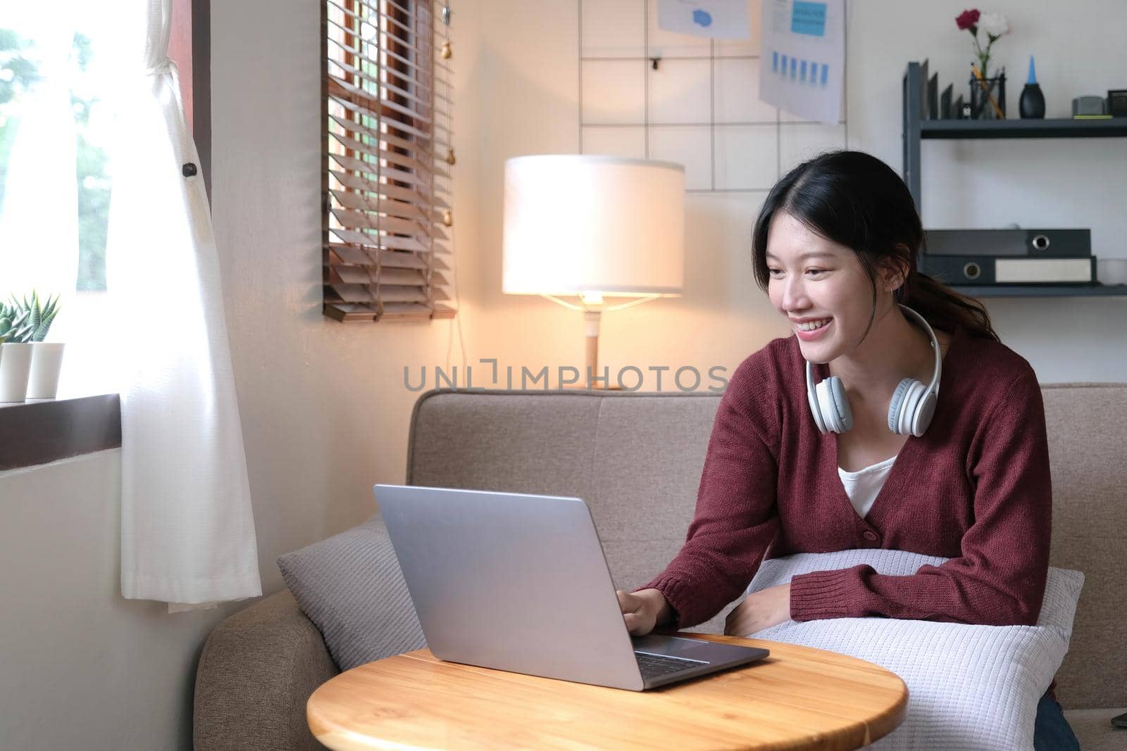 Beautiful Asian woman using laptop while headphones sitting on sofa at home by wichayada