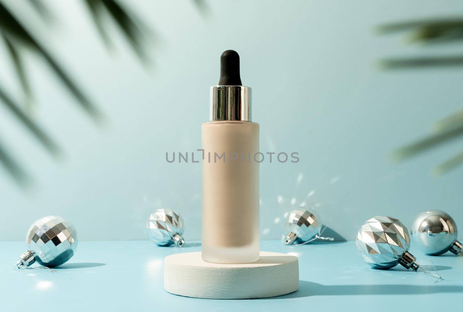 A minimalistic scene of foundation dropper bottle on podium with christmas decorative balls and pine tree on light blue background by Desperada