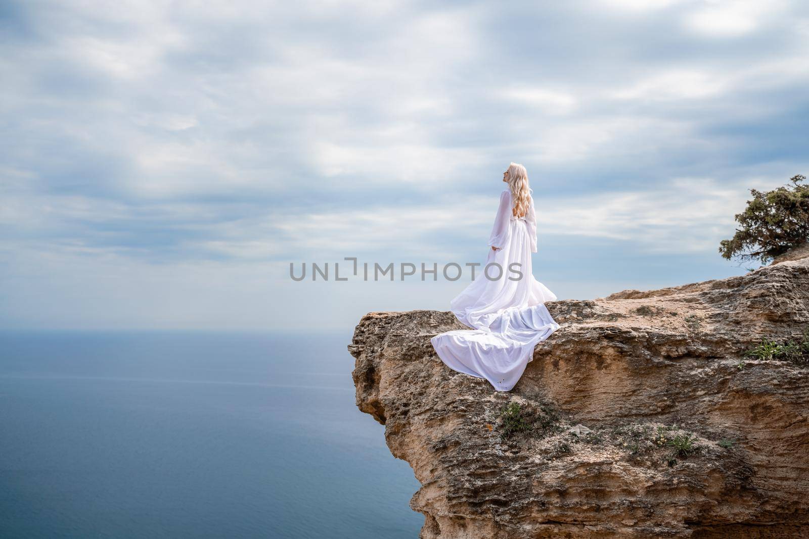 Blonde with long hair on a sunny seashore in a white flowing dress, rear view, silk fabric waving in the wind. Against the backdrop of the blue sky and mountains on the seashore