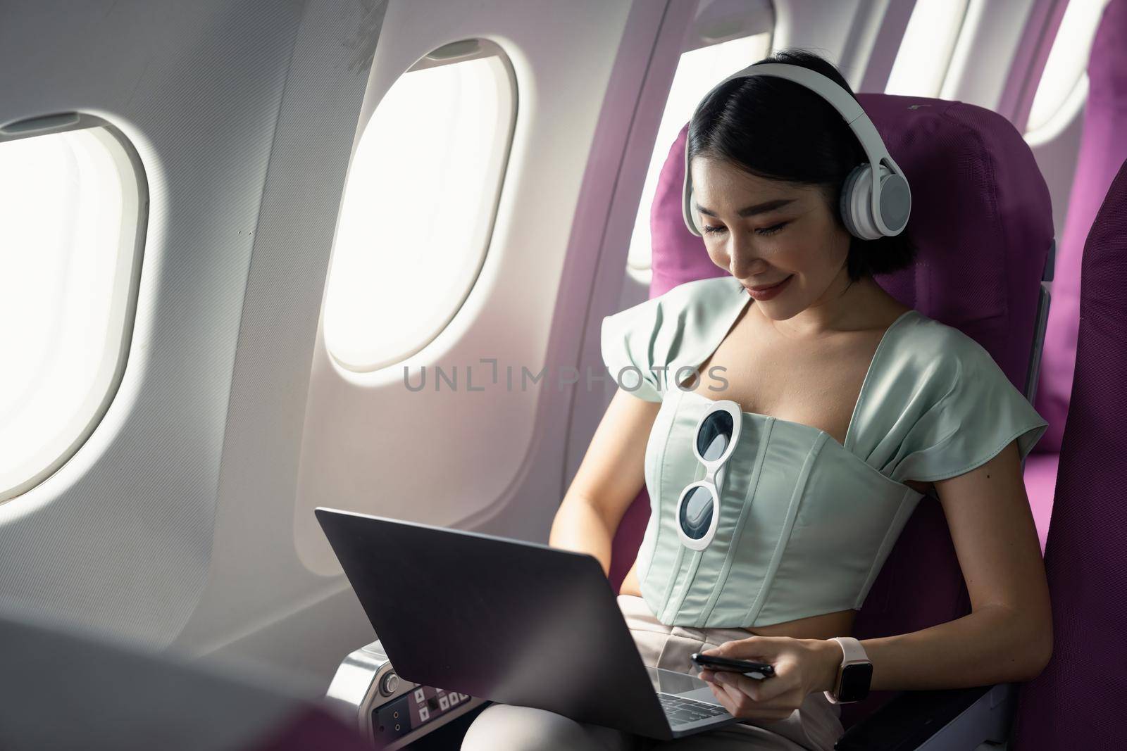 Plane passenger asian business woman working in airplane cabin during flight with laptop computer wifi listening to music with headphones by nateemee