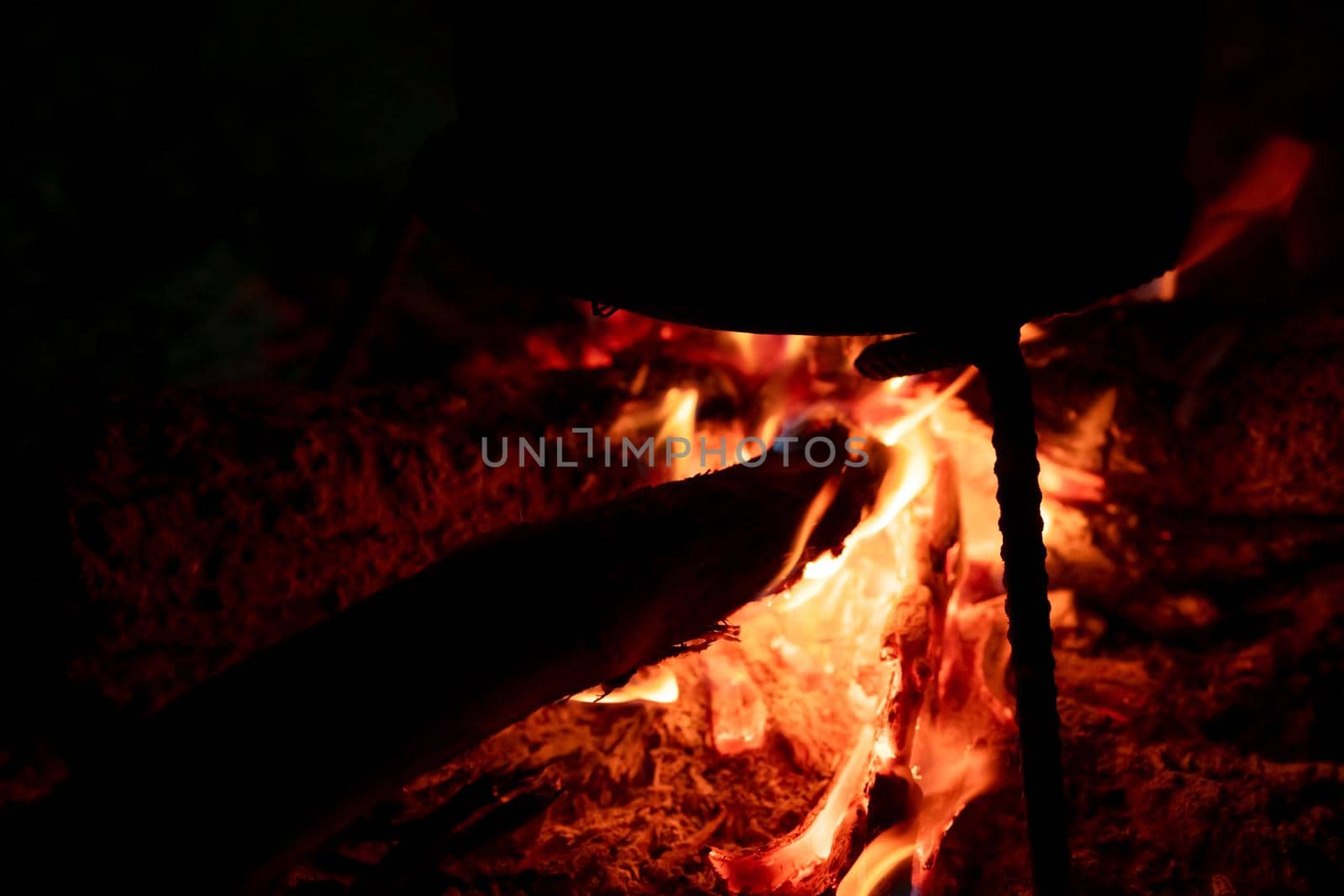 Food cooking on a bonfire at night. Firewood solid fuel for cook at campfire. Fire with orange flame on dark background. Burning wood for heating energy. Dinner at camping. Fire to keep warm at camp.