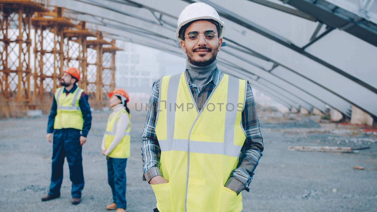 portrait of Arab man wearing safety helmet standing in construction site smiling looking at camera while people in uniform working in background