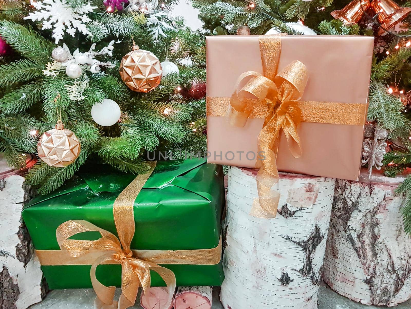 Beautiful boxes wrapped in gold paper with Christmas gifts on a birch stump near the Christmas tree in the room.