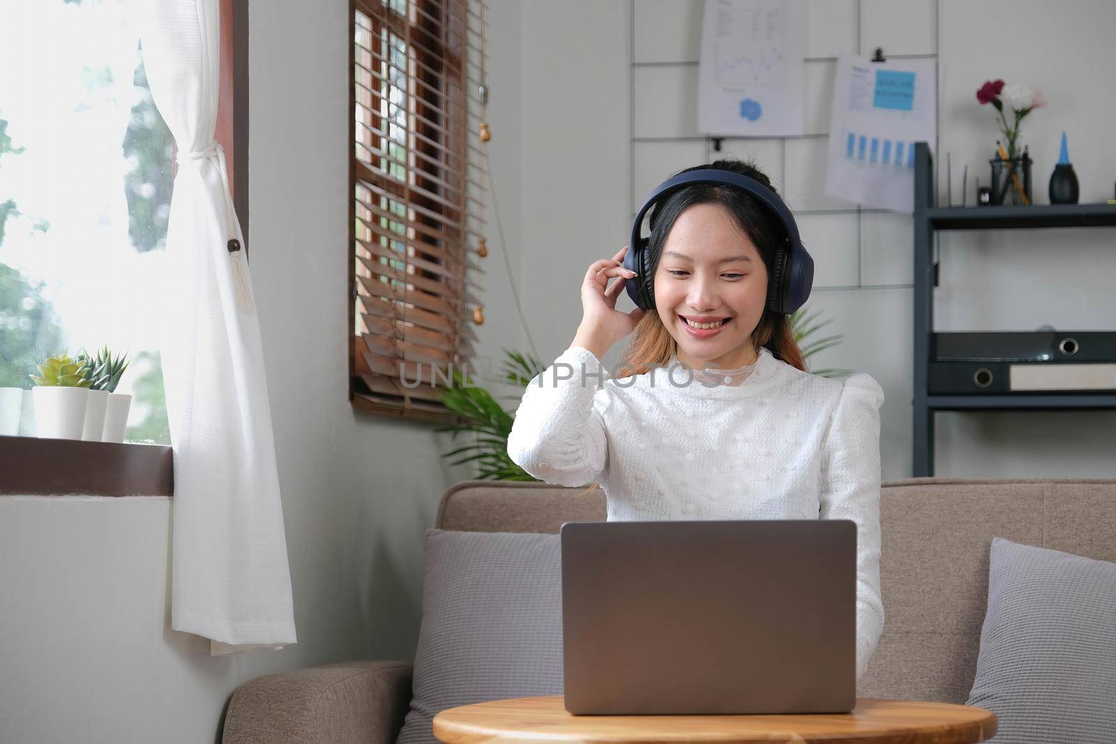Young adult happy smiling Hispanic Asian student wearing headphones talking on online chat meeting using laptop in university campus or at virtual office. College female student learning remotely. by wichayada