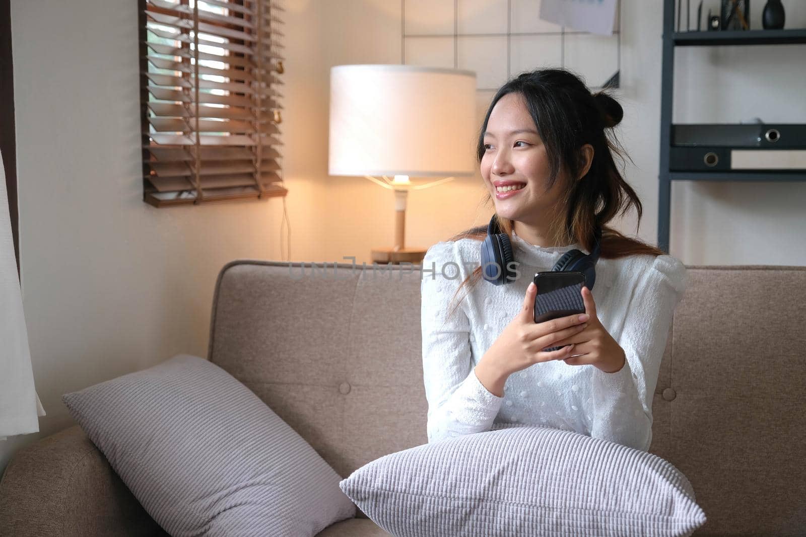 Happy and cheerful young Asian female choosing her music playlist on mobile phone, listening to music through her headphones while relaxing in the living room..