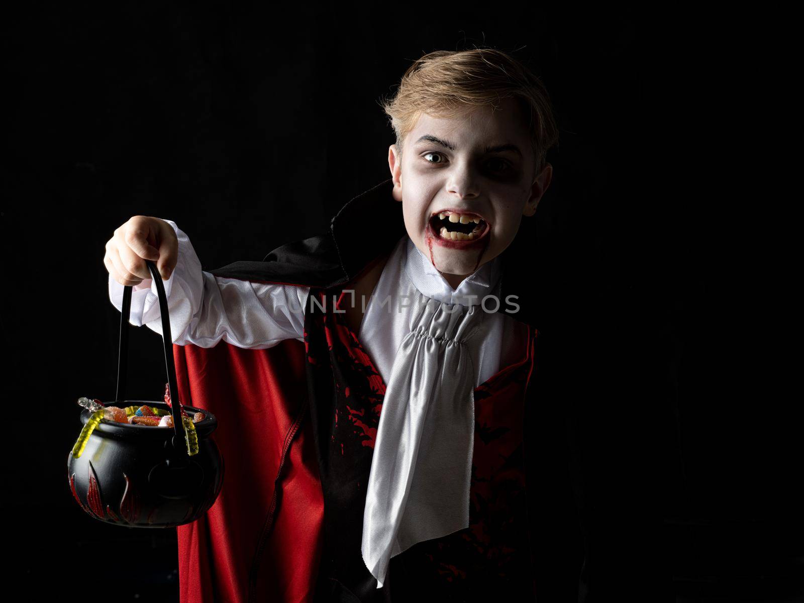 Happy child boy in costume of vampire holding cauldron od candies on black background in holiday Halloween