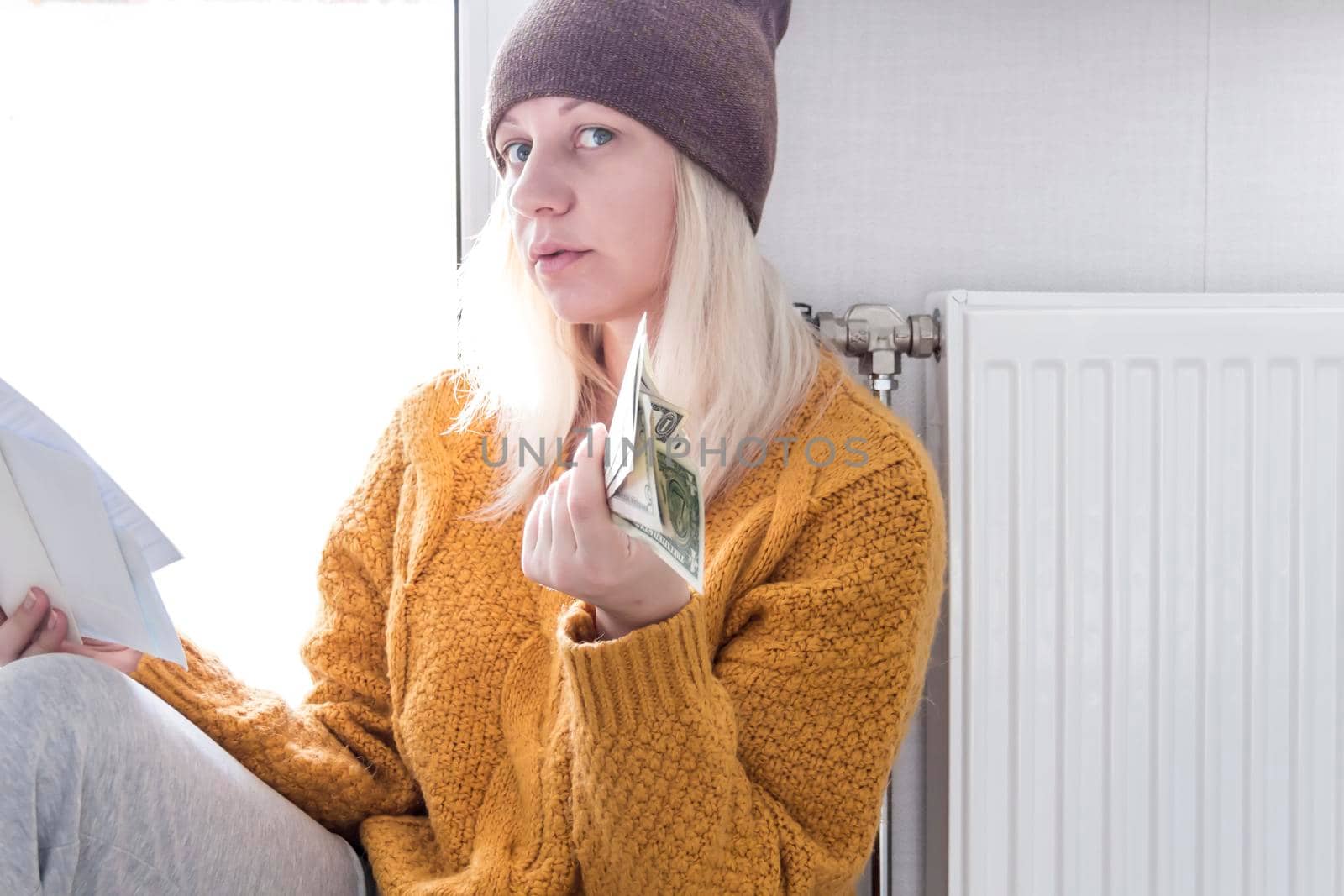 A young girl in a yellow sweater and a brown hat is sitting on the floor, counting money and thinking how to pay bills and taxes near a heater with a thermostat..