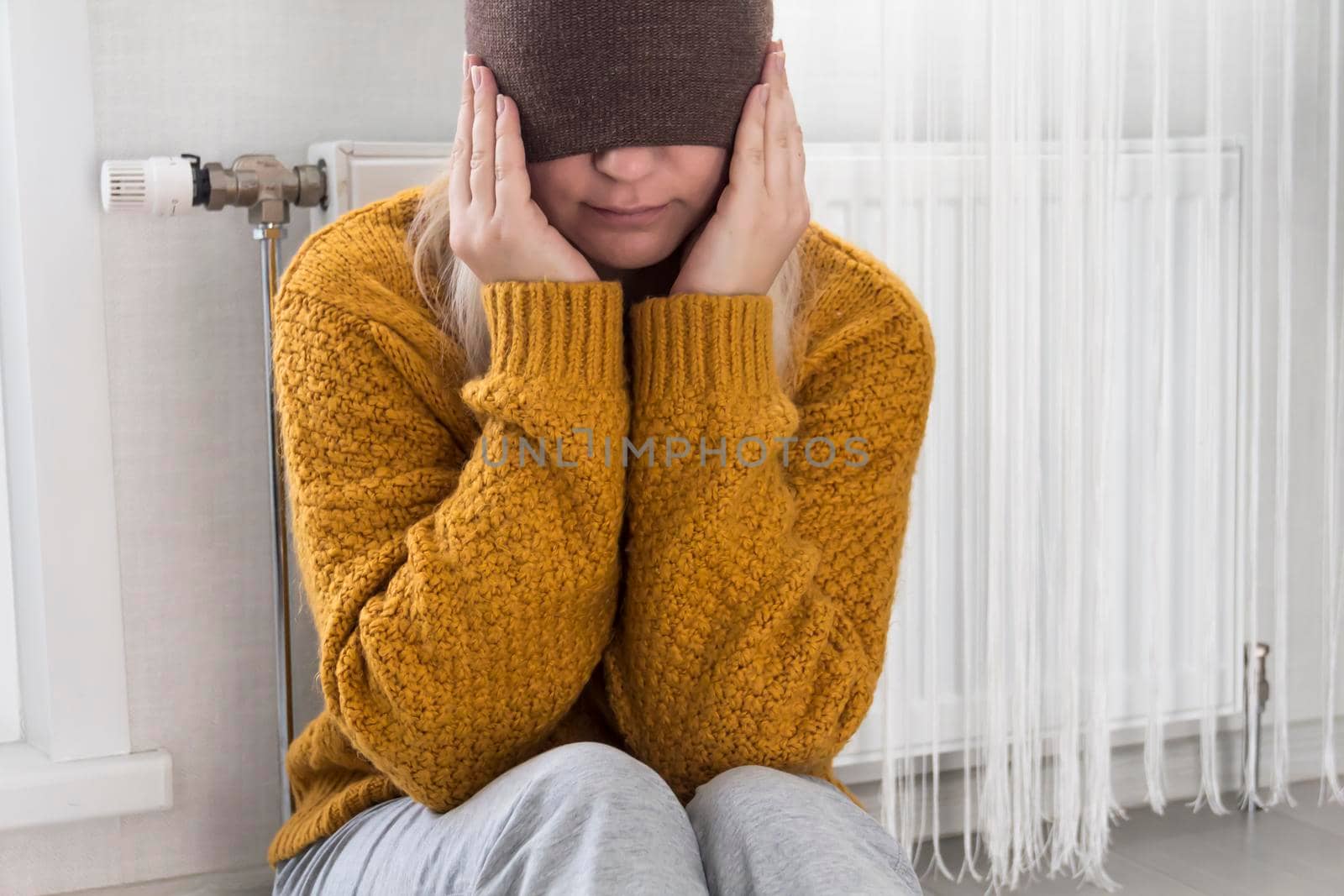 A young girl in a yellow sweater is sitting on the floor, pulling a brown hat over her face near a heater with a thermostat..