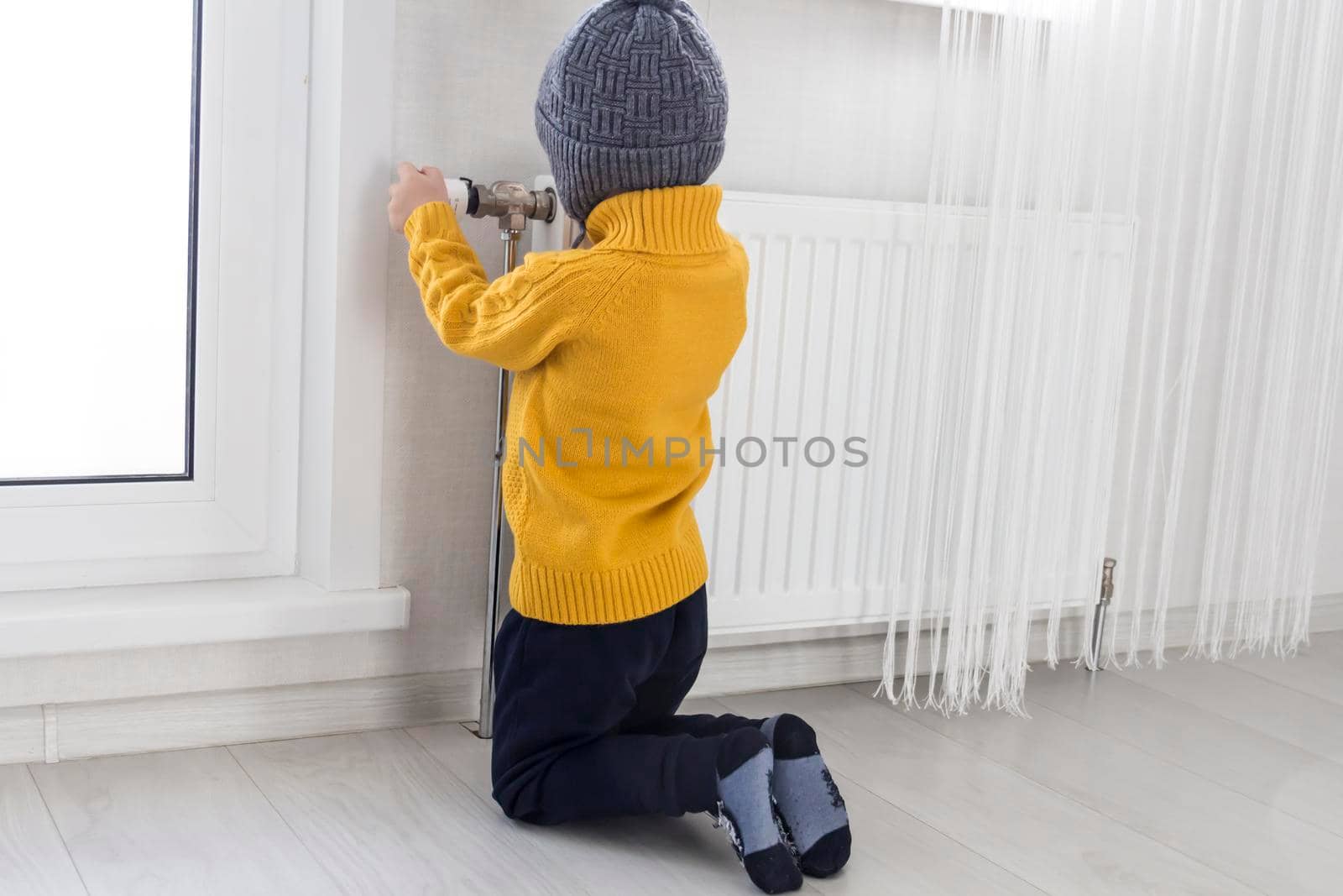 A small child in a yellow sweater and a gray hat is kneeling near a heater with a thermostat...