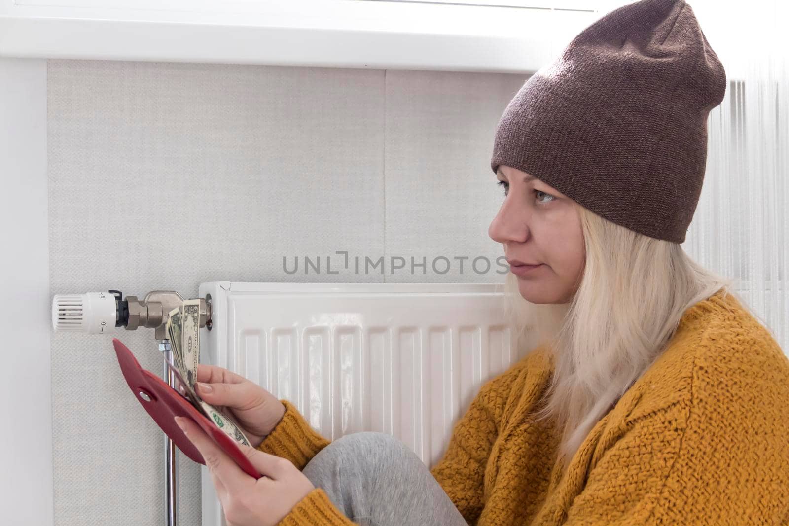 A young girl in a yellow sweater and a brown hat is sitting on the floor, counting money and thinking how to pay bills and taxes near a heater with a thermostat..