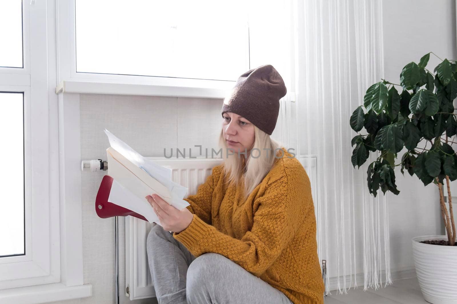 A young girl in a yellow sweater and a brown hat is sitting on the floor, counting money and thinking how to pay bills and taxes near a heater with a thermostat..