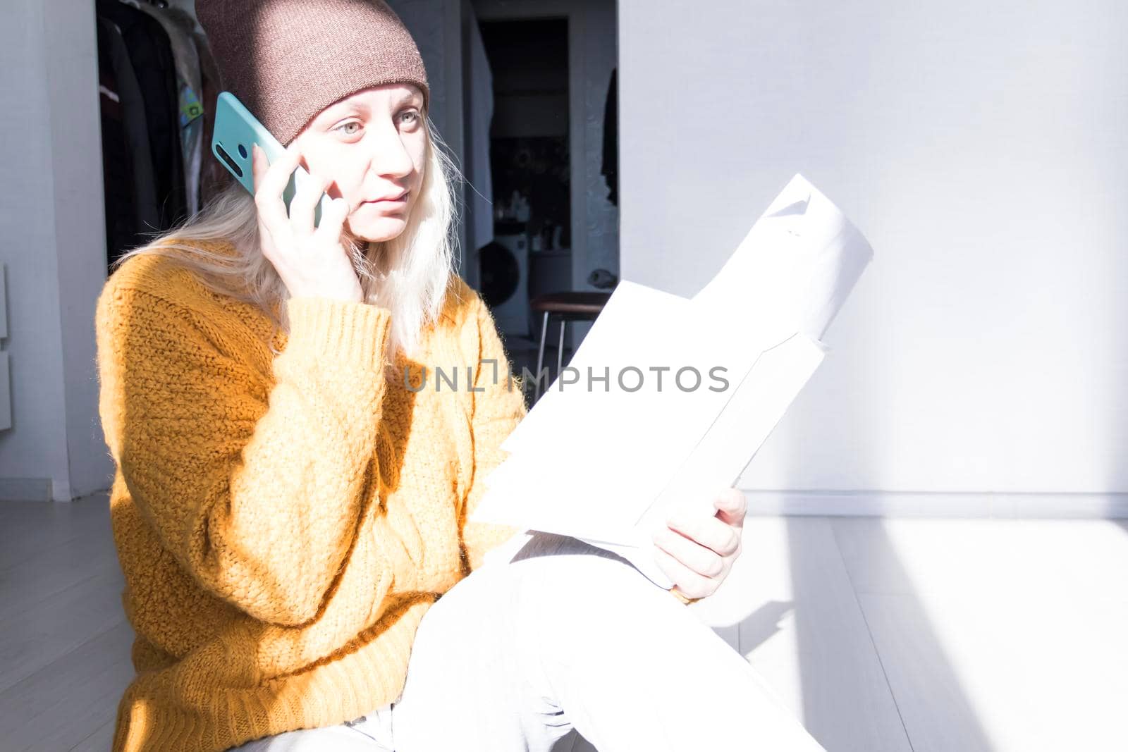 A young girl in a yellow sweater and hat is talking on a mobile phone and studying large bills and utility bills..