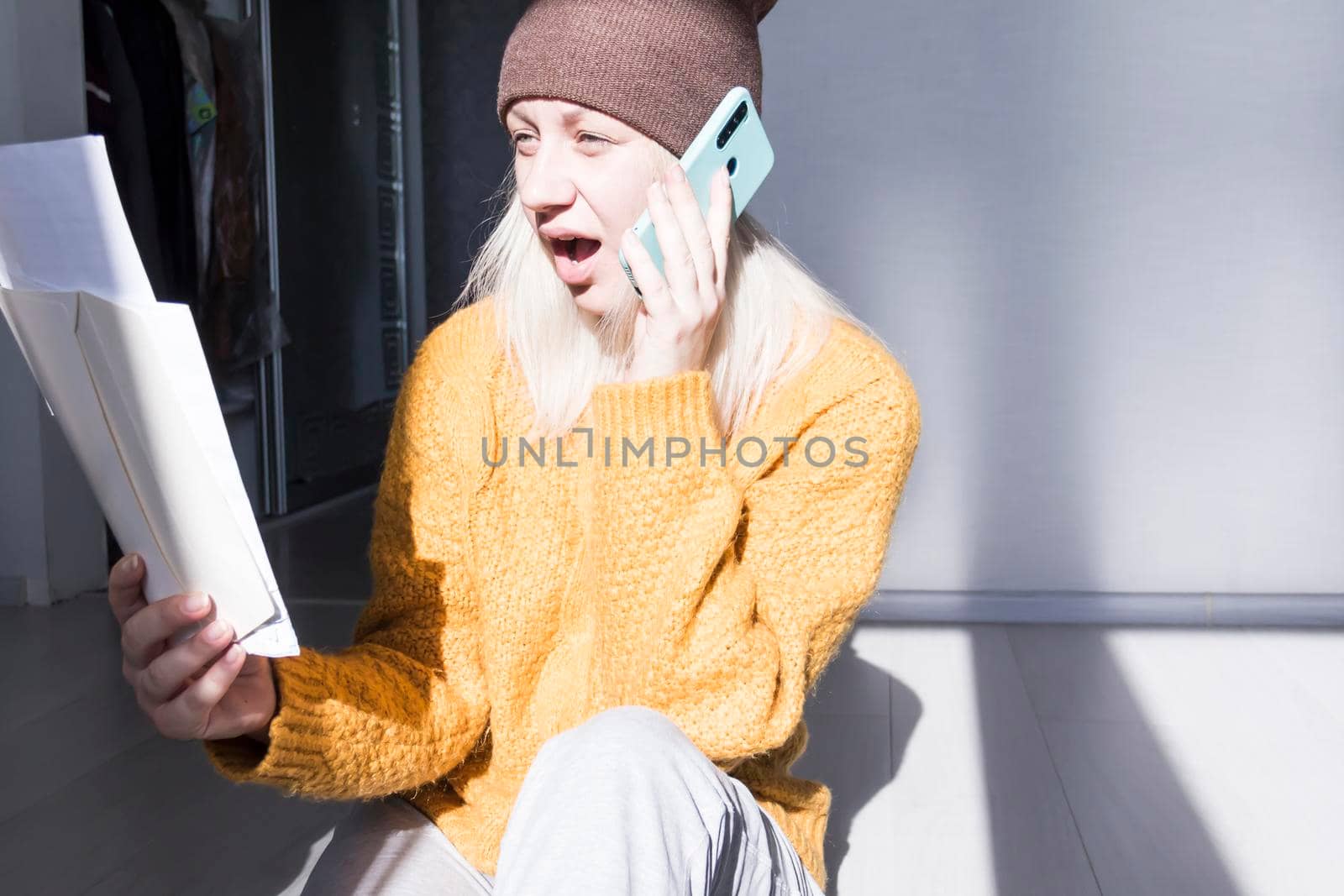A young girl in a yellow sweater and hat is talking on a mobile phone and studying large bills and utility bills..