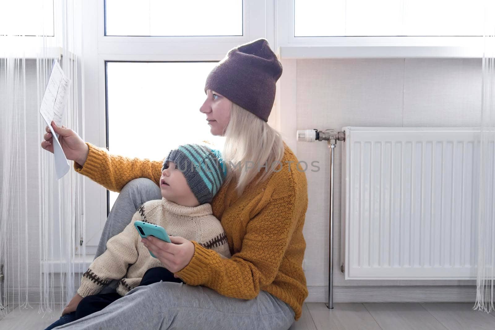 .A mother with a small child in a yellow sweater and hats is counting money and thinking how to pay bills and taxes, near a heater with a thermostat.