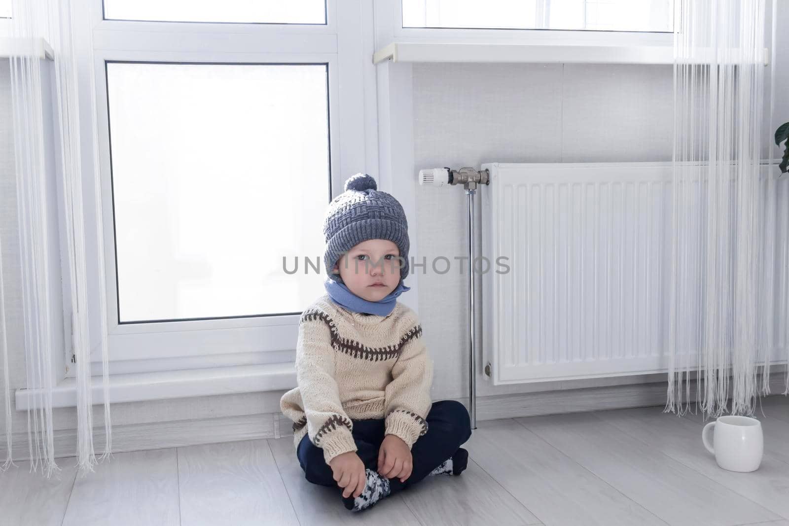 A small child in a sweater and a gray hat is kneeling near a heater with a thermostat...