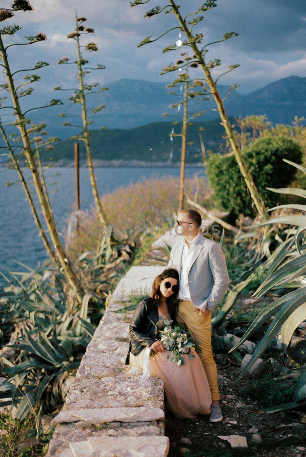 Bride sitting on a stone fence leaned against the standing groom. High quality photo