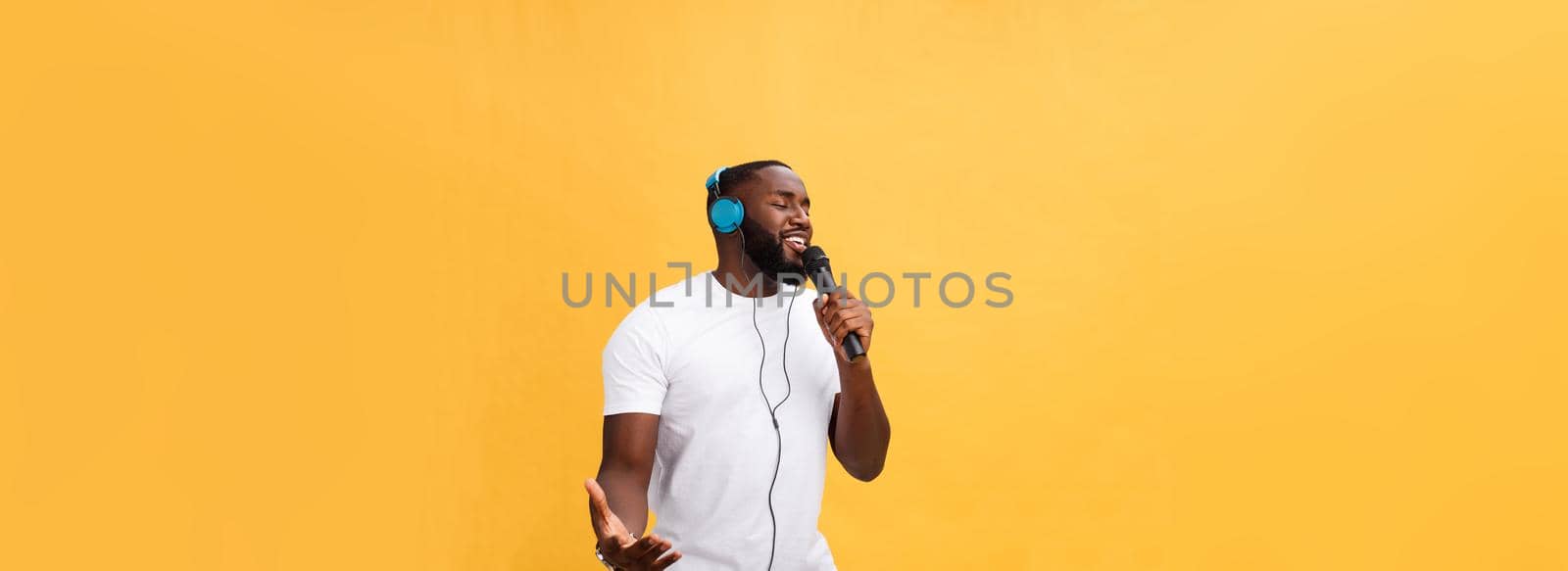 Portrait of cheerful positive chic. handsome african man holding microphone and having headphones on head listening music singing song enjoying weekend vacation isolated on yellow background.