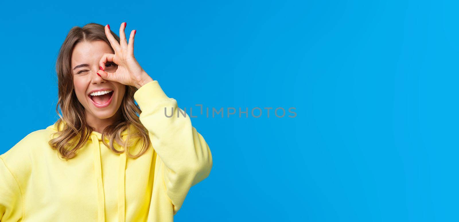 Close-up portrait of optimistic dreamy caucasian girl with blonde short hair, showing okay gesture over eye and wink happy, express positive enthusiastic emotion, standing blue background by Benzoix