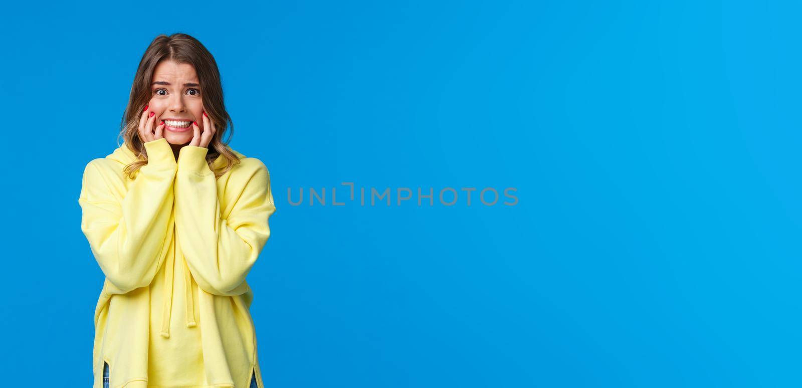Embarrassed and afraid young timid blond caucasian female in yellow hoodie panicking, biting fingernails from fear and worries, looking scared with alarmed expression, blue background by Benzoix