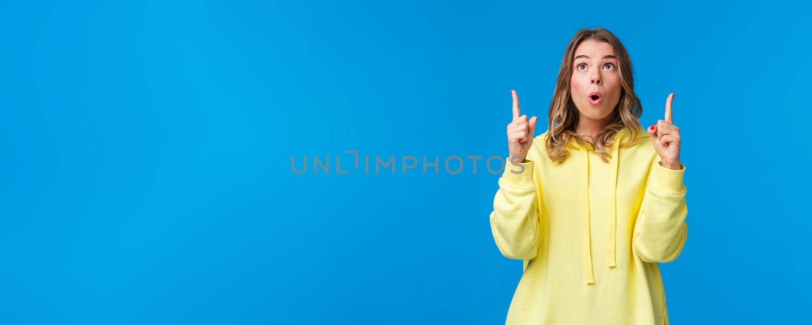 Dreamy and impressed startled cute caucasian female in yellow hoodie, make surprised and curious face with lips folded in wow, look and pointing up, top advertisement, blue background by Benzoix