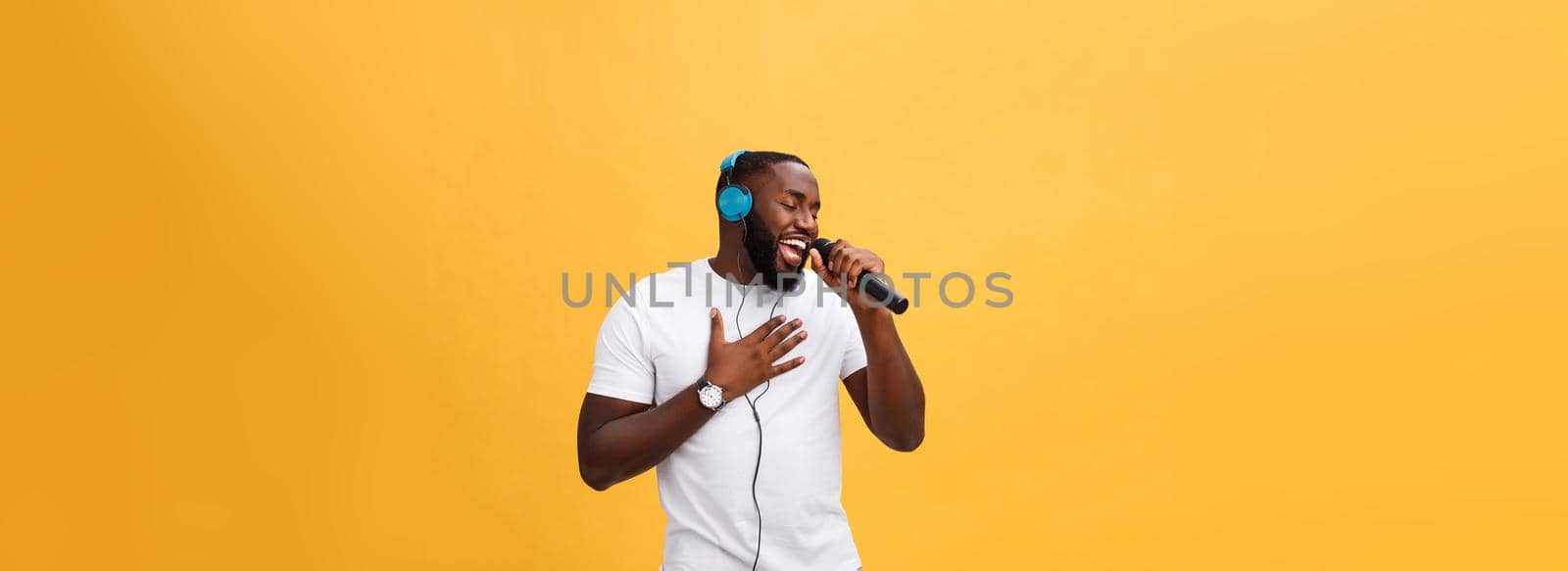 Portrait of cheerful positive chic. handsome african man holding microphone and having headphones on head listening music singing song enjoying weekend vacation isolated on yellow background.