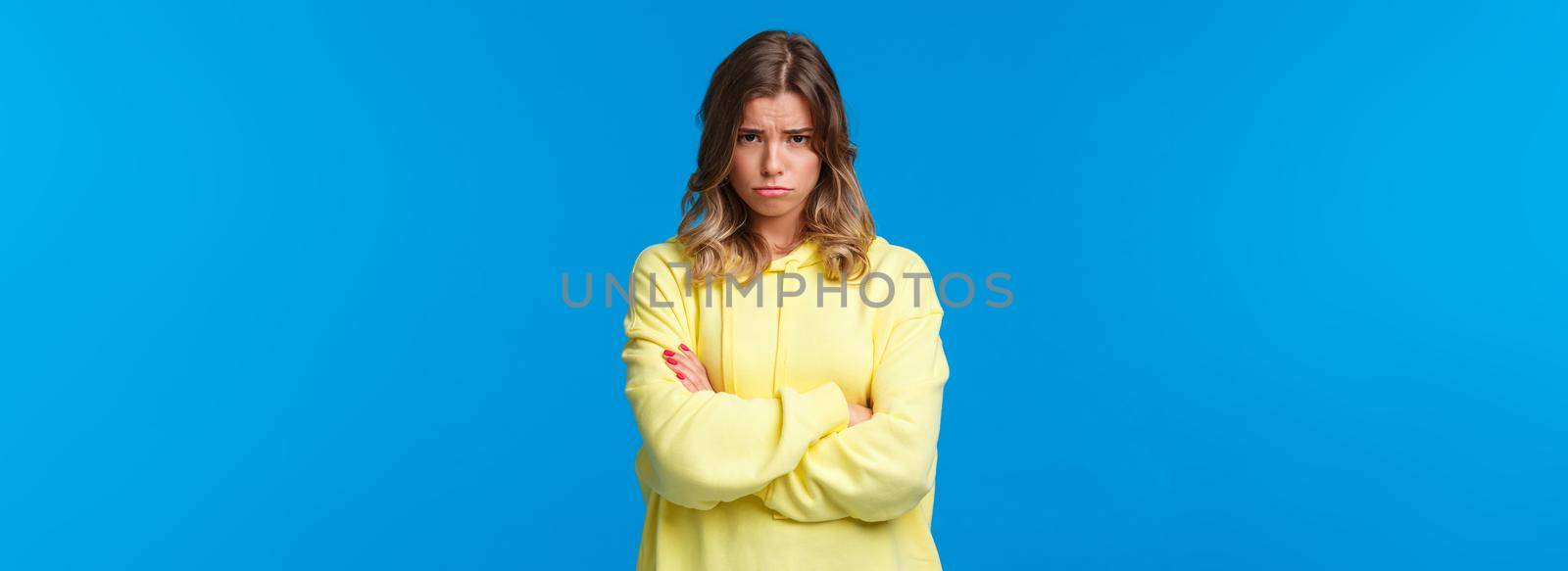 Gloomy and moody cute blond teenage girl look from under forehead and sulking at camera, feeling offended or upset, cross hands chest insulted waiting apology, blue background by Benzoix
