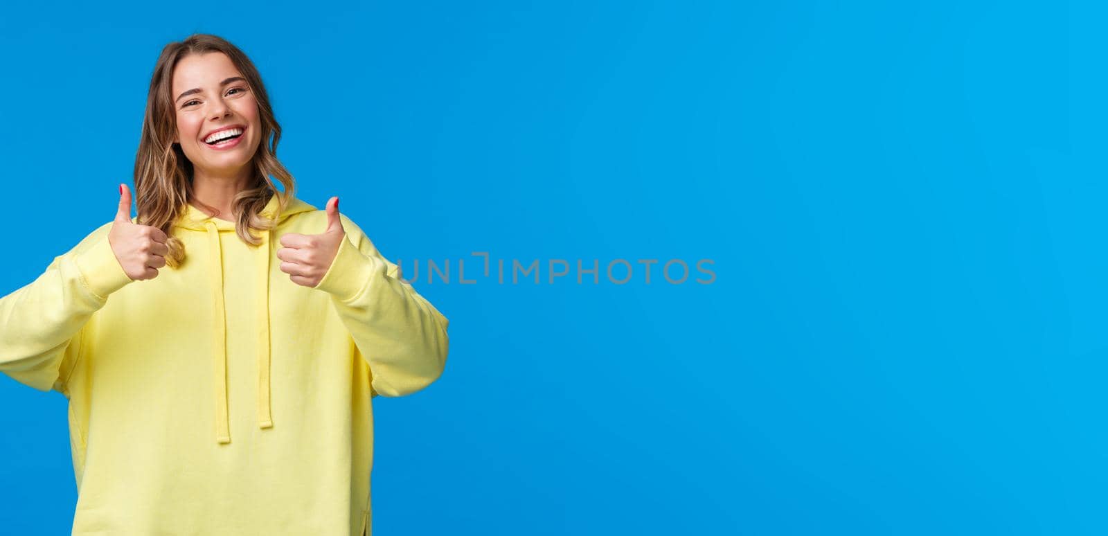 Sounds good Im in. Cheerful and optimistic good-looking young female blonde in yellow hoodie, show thumbs-up in approval, smiling nod acceptingly, like idea, blue background.