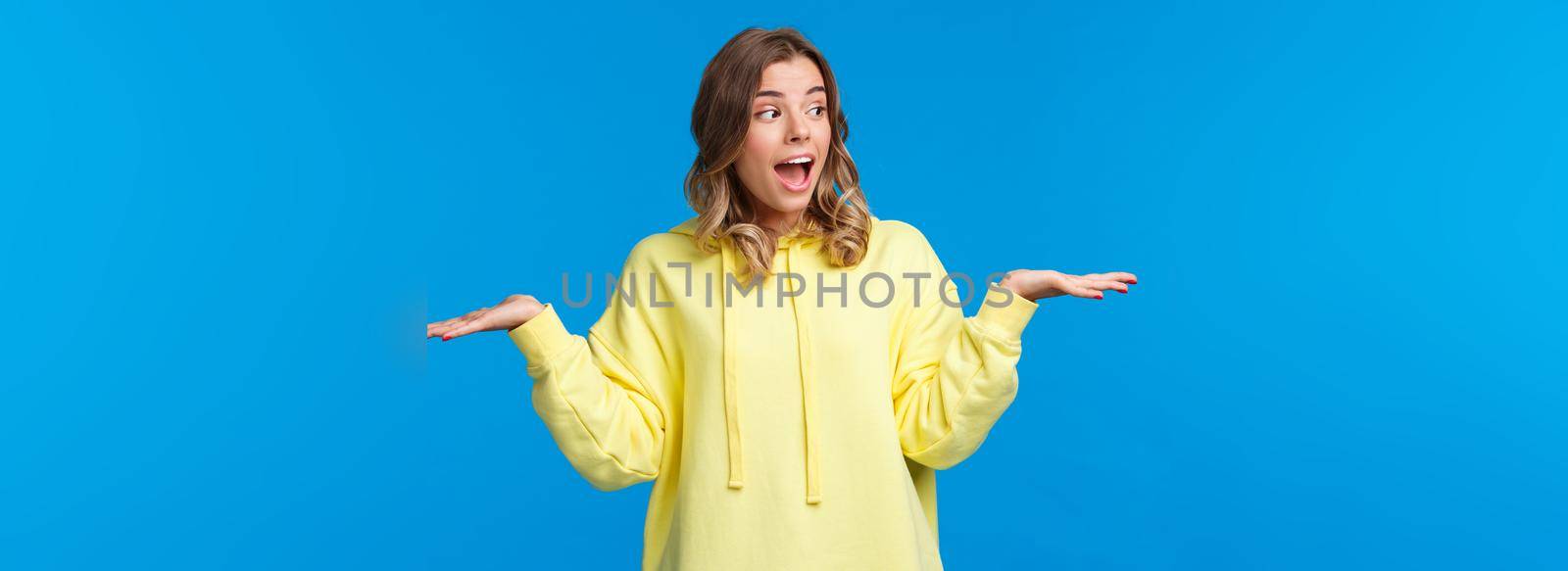 Girl weighing two choices as making decision, look away picking between products she holding in arms on left and right side, standing excited over blue background.