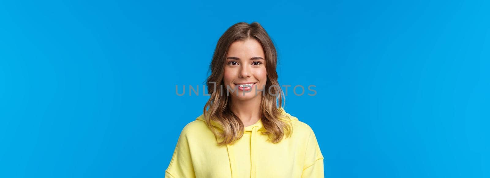 Close-up portrait gorgeous blond caucasian young girl with short haircut, wearing yellow hoodie, smiling with beaming white grin, looking camera, standing blue background. Copy space