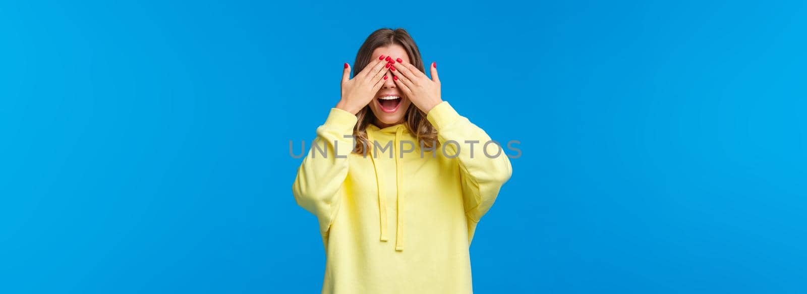 Girl waiting for friends tell her open eyes as shut it with hands and count ten during b-day party, girlfriend bring surprise gift, smiling with excitement, playing peekaboo, stand blue background.