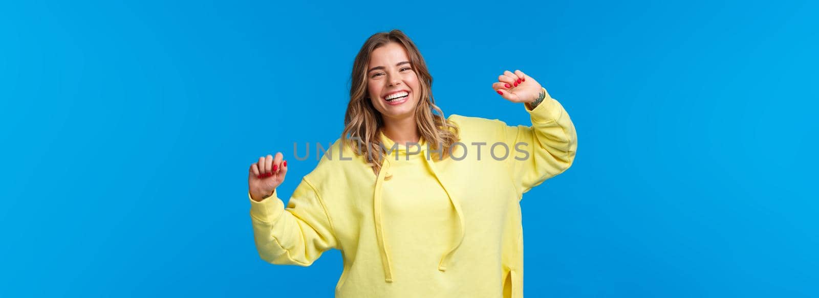 Carefree happy alluring young blond woman having fun, vibing on weekends, enjoying party as dancing and laughing over blue background, lift hands up joyful by Benzoix