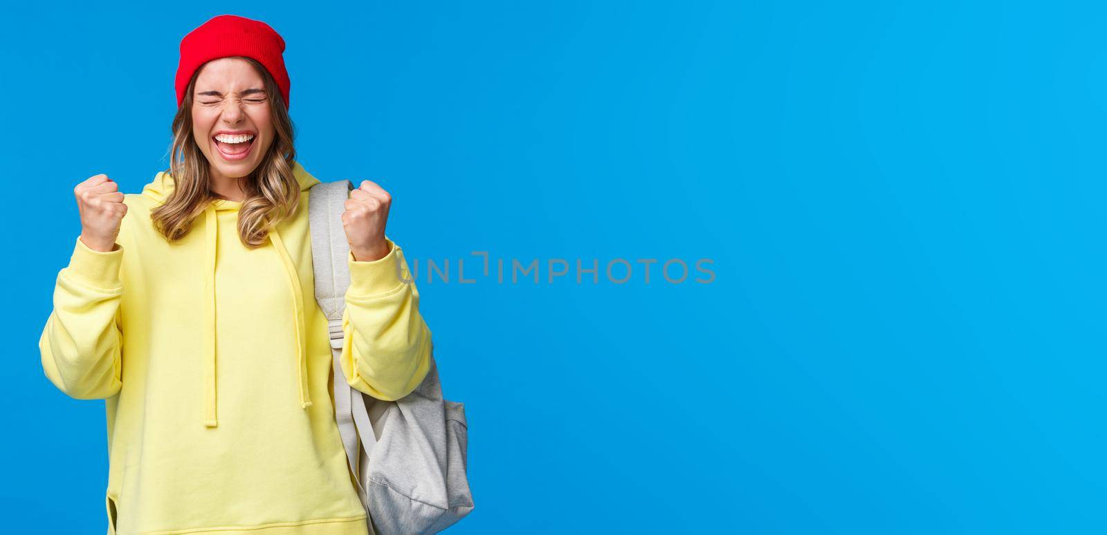 Happy cheerful blond girl in red beanie and yellow hoodie, close eyes relieved and smiling triumphing, celebrating great news, passed exams in university, fist pump like champion, winning prize by Benzoix