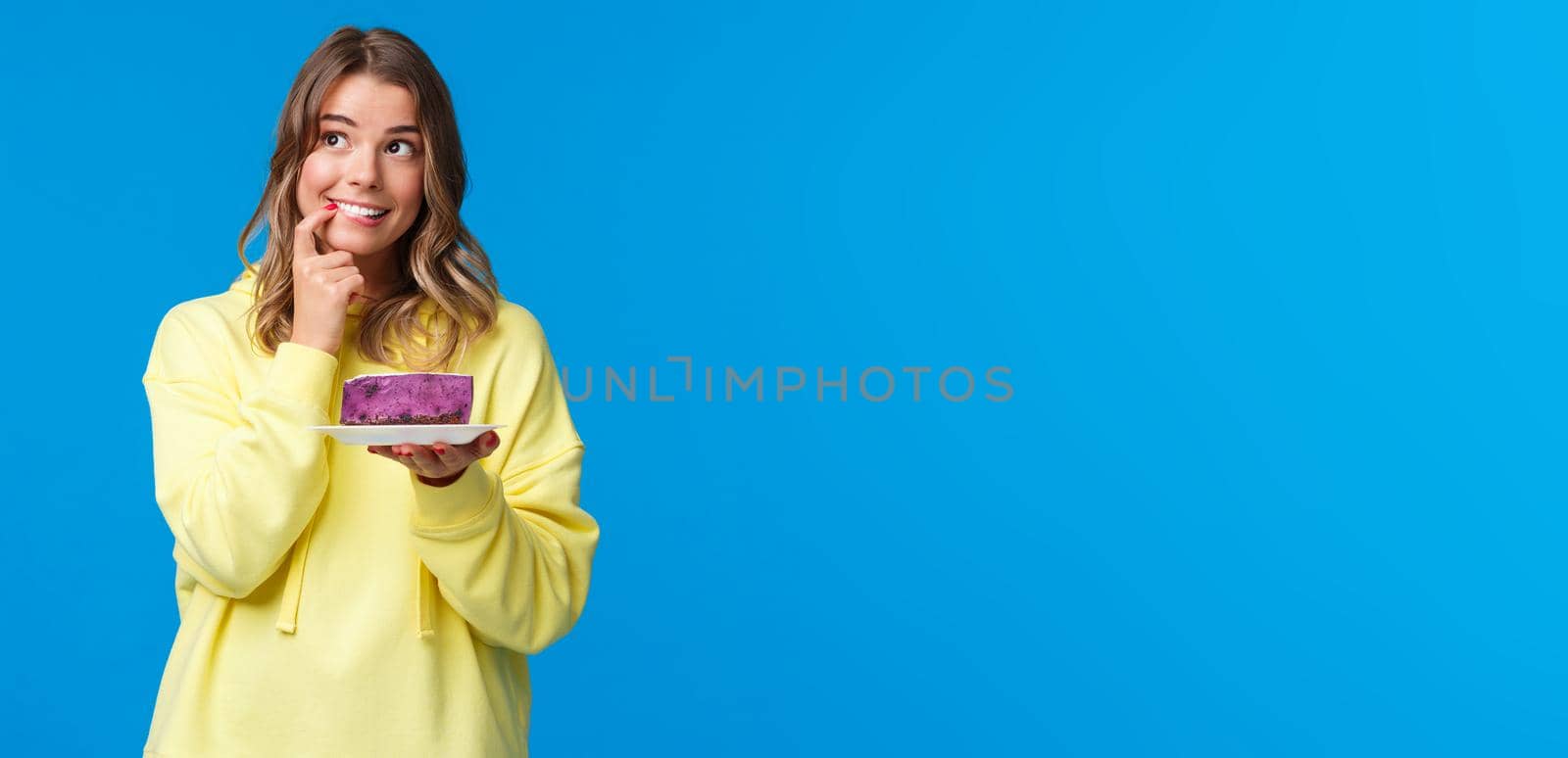 Celebration, party and lifestyle concept. Dreamy cute and silly blond girl thinking what to wish, biting finger and looking thoughtful up, smiling holding b-day cake, blue background by Benzoix