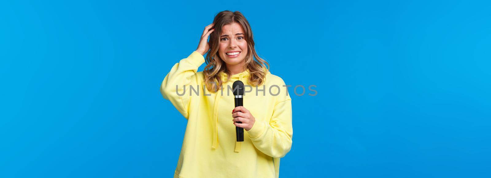 Embarrassed girl holding microphone awkward smiling and scratch head as singing song on karaoke with friends, shy perform in front of audience, standing blue background.