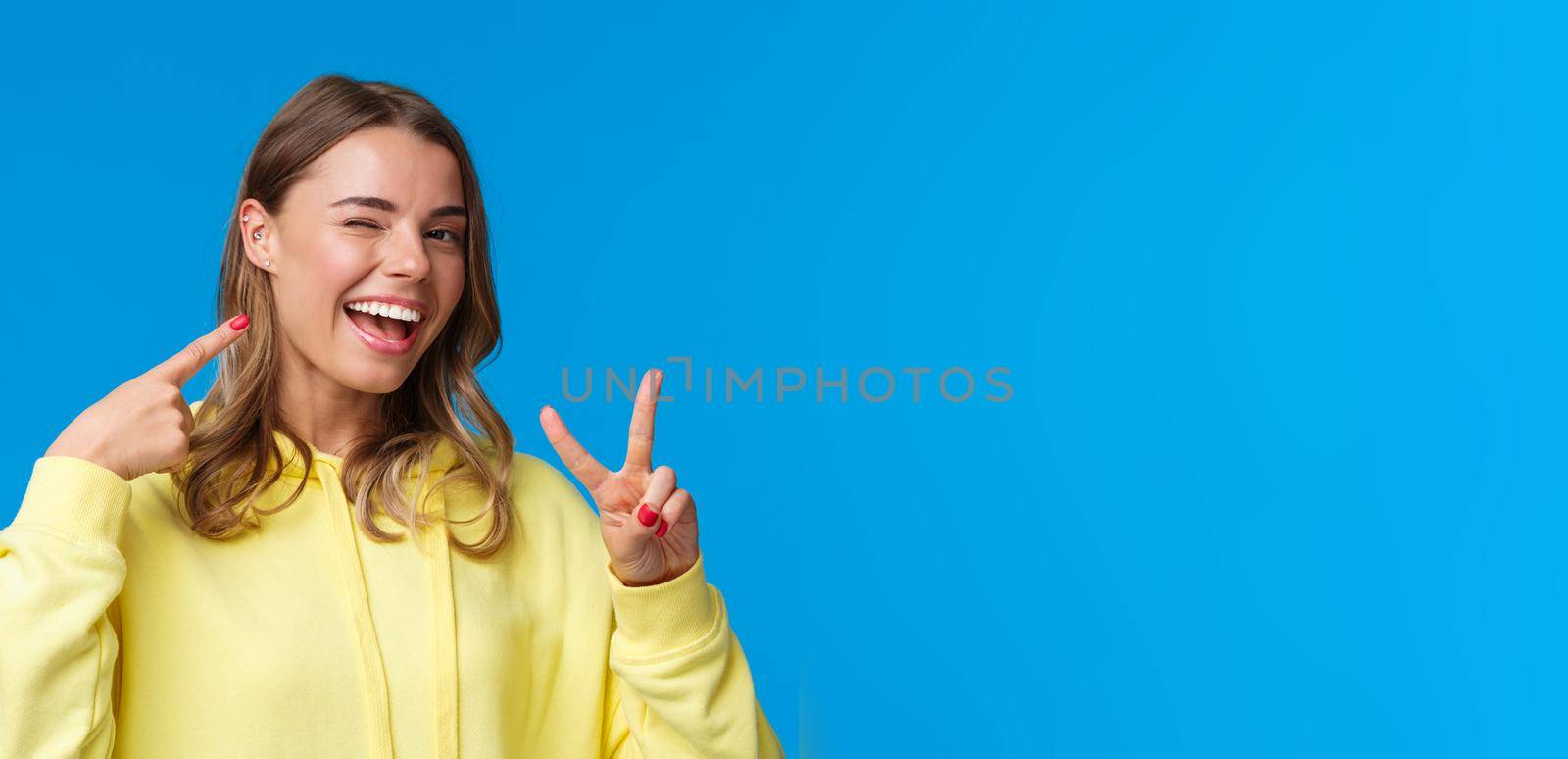Close-up portrait of proud and boastful cute blond girl with short hair and pierced ear, pointing at herself and make peace gesture with pleased smile, standing blue background upbeat.