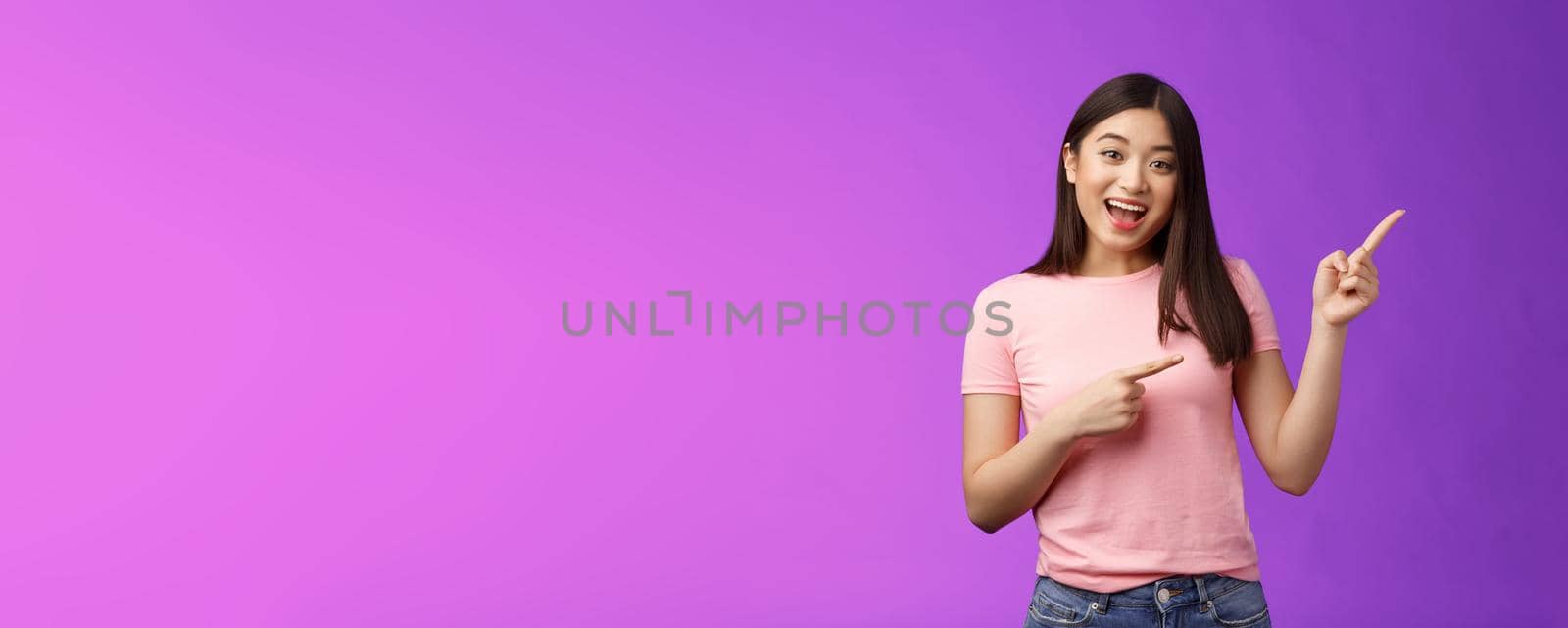 Friendly enthsuasitic amazed attractive tender asian girl having interesting conversation, discuss interest new store, pointing right inviting come inside, stand purple background smiling camera.