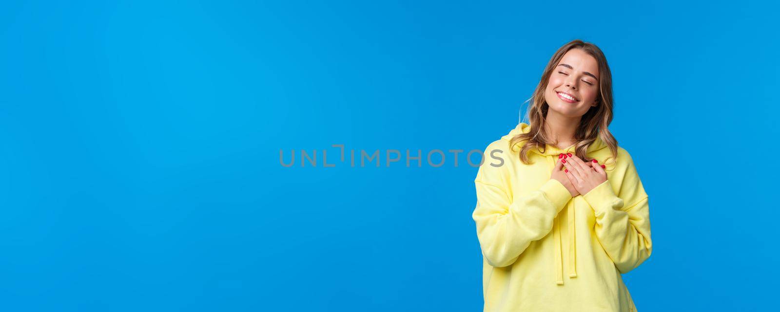 Romantic alluring blond caucasian girlfriend in yellow hoodie, press hands to heart and close eyes with lovely smile recall beautiful memory, imaging perfect date, standing blue background by Benzoix