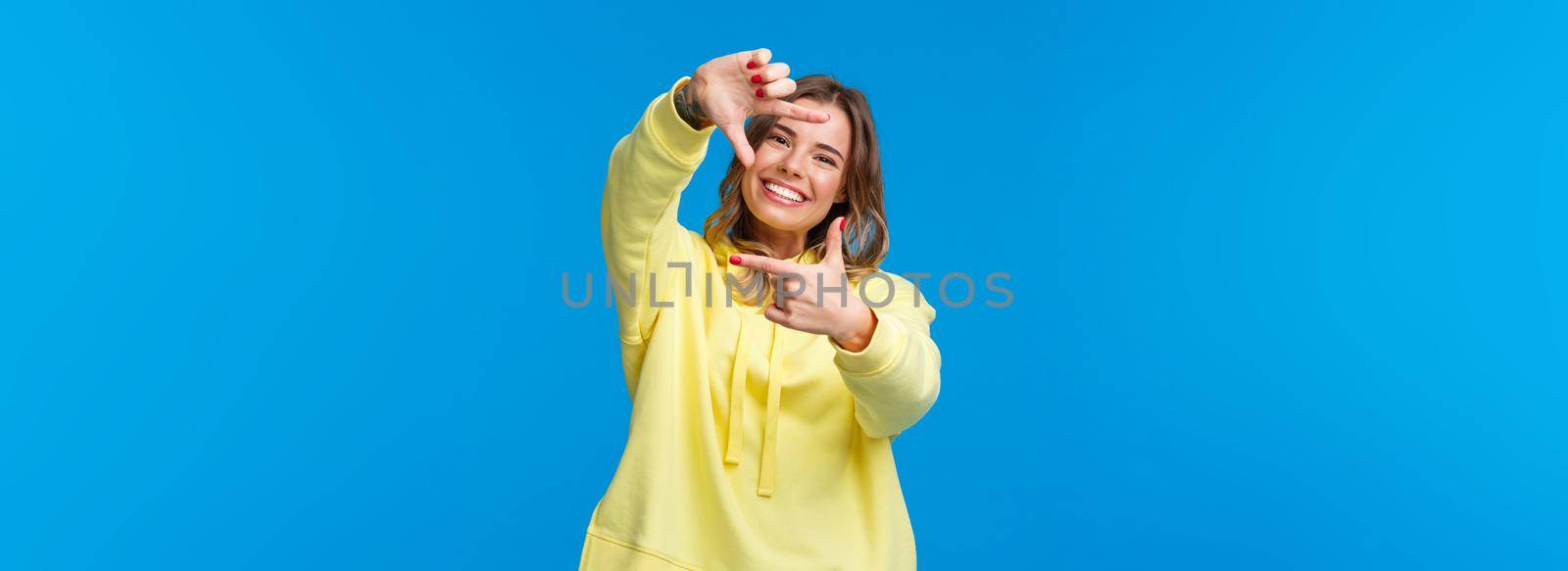 Inspiration, lifestyle and people concept. Portrait of cheerful creative blond girl searching perfect angle for shot, look at camera through frame gesture and smiling, blue background.