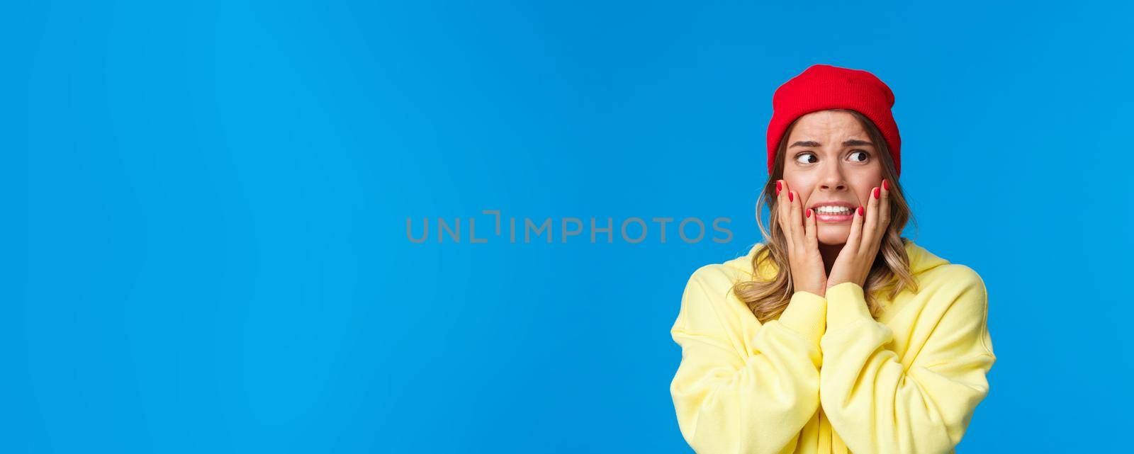 What have I done. Worried and concerned anxious caucasian female in red beanie and yellow hoodie, hold hands on cheeks panicking look away frightened, standing troubled blue background by Benzoix