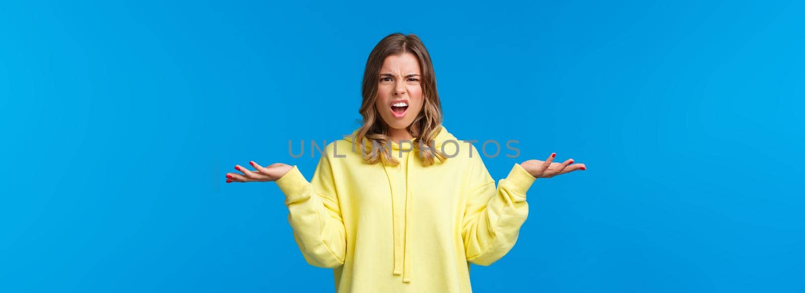 What problem wtf. Pissed-off frustrated and angry young blond caucasian girl shrugging with confused annoyed expression, cant understand what happened, dont know anything, blue background.